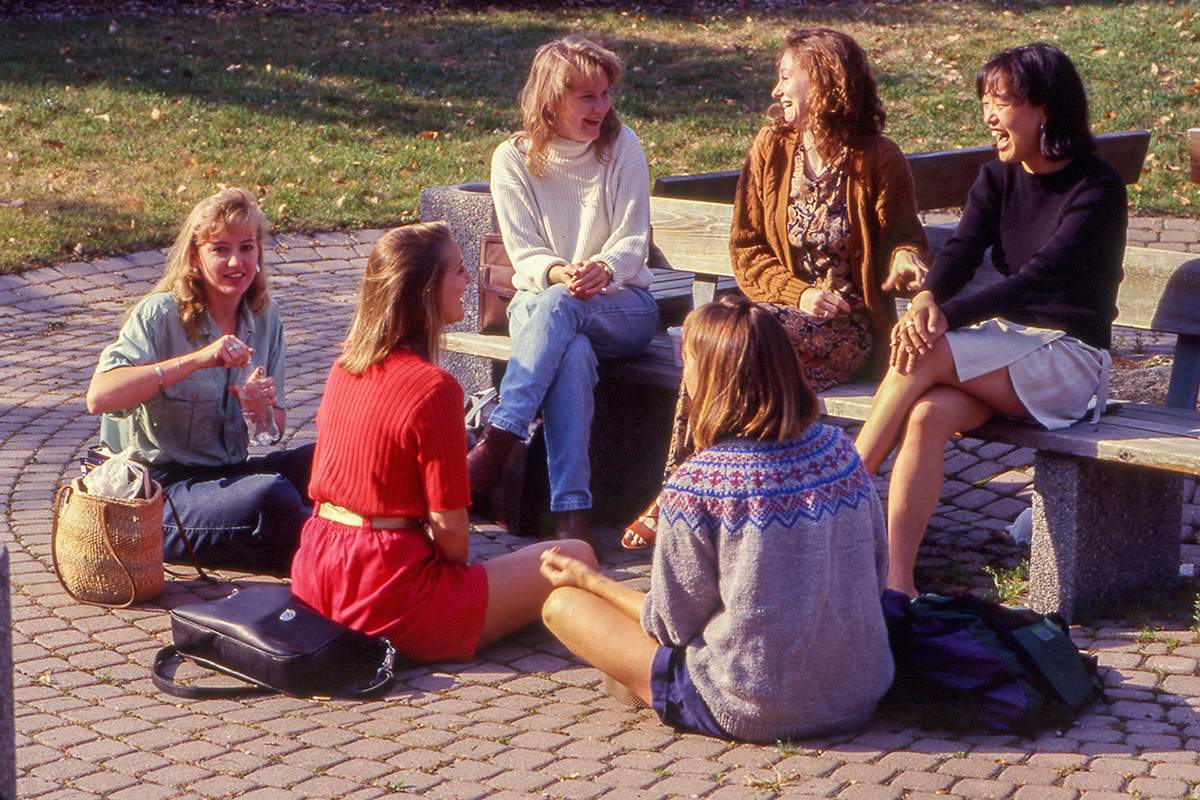 A group of young women share a laugh as they sit on and stand around a park bench