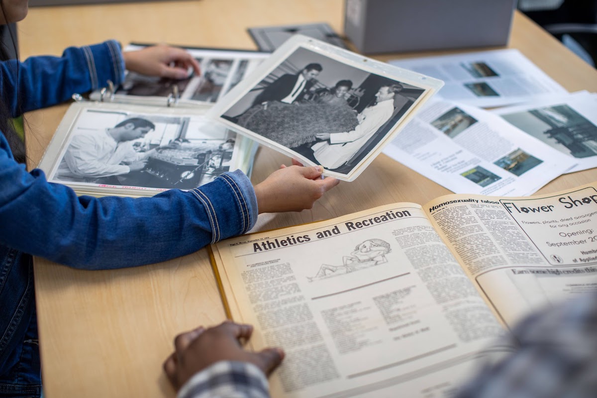 Students leafing through archival items scattered on a desk, including black-and-white photographs and a yellowed publication.