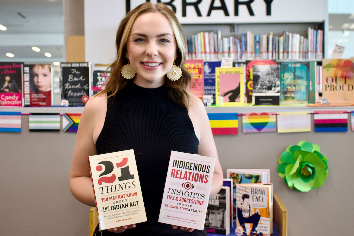 A smiling team member in front of a Library display, holding up two books: “21 Things You May Not Know about the Indian Act” by Bob Joseph and “Indigenous Relations” by Bob Joseph and Cynthia F. Joseph.