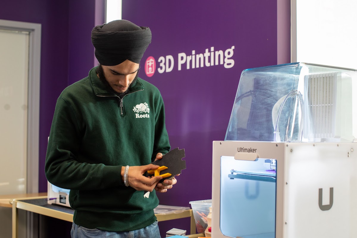 A student standing in front of an Ultimaker 3D printer, looking down and fiddling with a small, angular 3D object.
