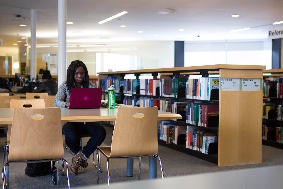 A student sitting at a table in the Library, working on a laptop. There are low bookshelves next to them and more students seated in the background.