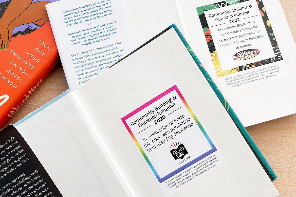 Two hardcover books open to show large stickers on the inside covers. One sticker has a rainbow border and reads: “Community Building & Outreach Initiative 2020. In celebration of Pride, this book was purchased from Glad Day Bookshop.” The other sticker has a colourful border and reads: “Community Building & Outreach Initiative 2022. To celebrate Black voices from Canada and beyond, this book was purchased from A Different Booklist bookshop in Toronto.”